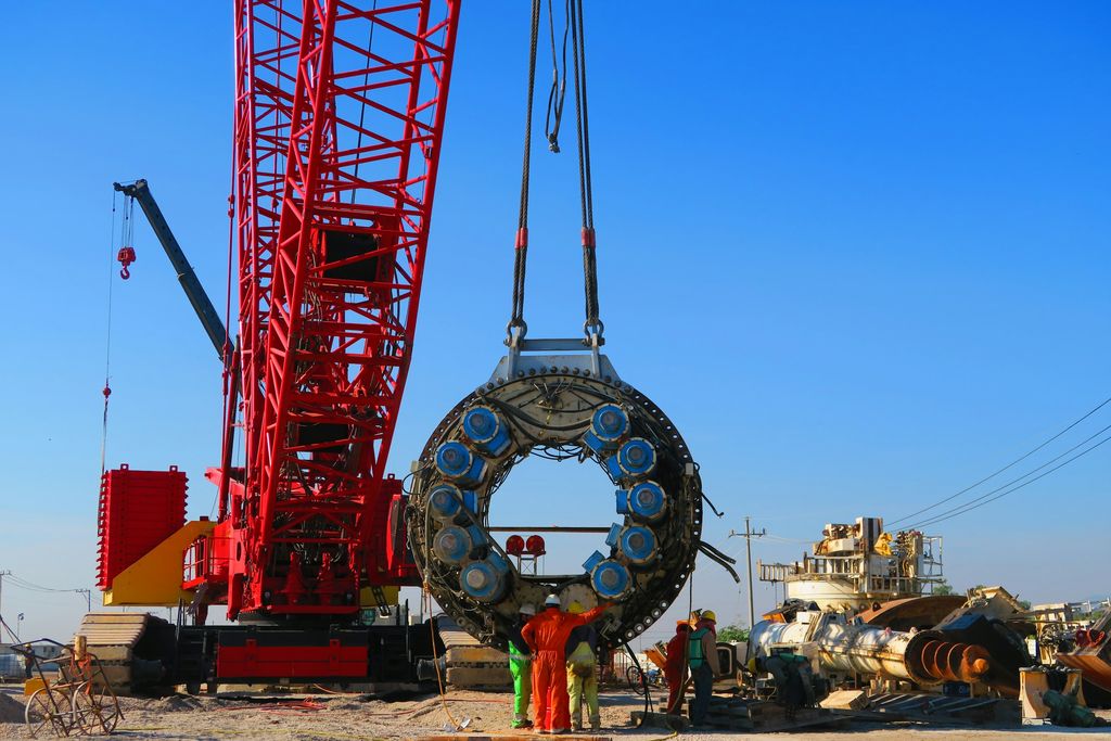 Red lattice-boom crawler crane rigging a large circular machine section while ground crew look on