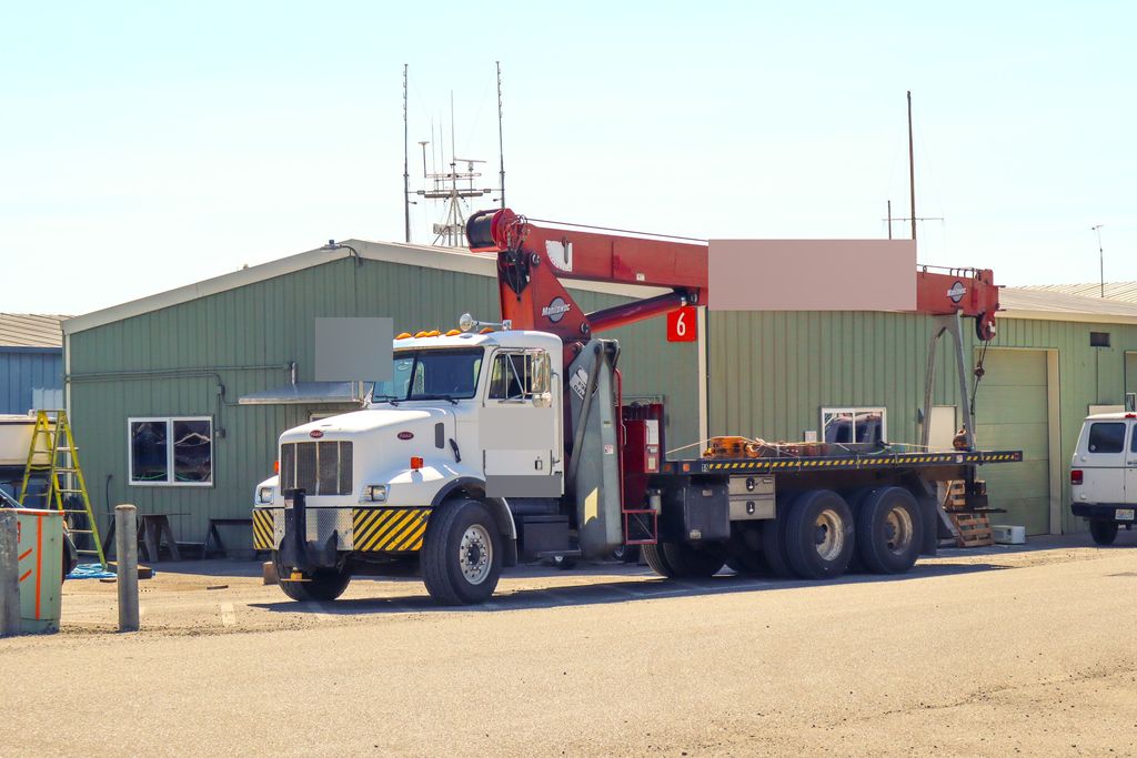 Truck-mounted crane with a red boom parked outside a metal shop building